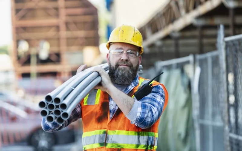 Construction worker carrying pipes on a job site — construction time tracking