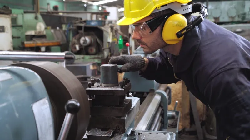 Machinist operating a lathe on a factory floor wearing hard hat and ear protection, manufacturing time tracking
