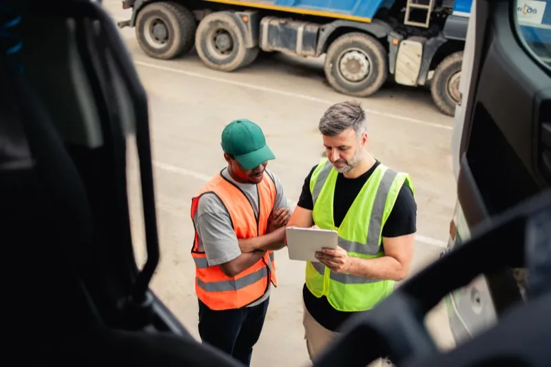 Two fleet drivers reviewing a tablet between trucks at a logistics yard — transportation time tracking